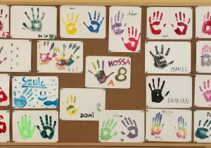 Wystawa prac uczniów klasy 8A. Na białych kartkach papieru znajdują się kolorowe odciski dłoni każdego z uczniów.